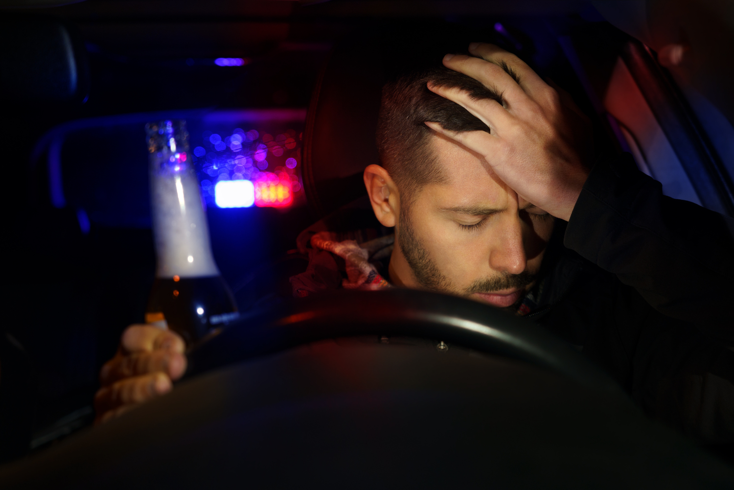 Man behind the wheel of a vehicle with hand on his head, blue and red police lights are visible through the read window.