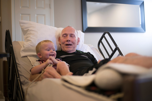 Elder man is sitting on bed with small toddler hugging and smiling.